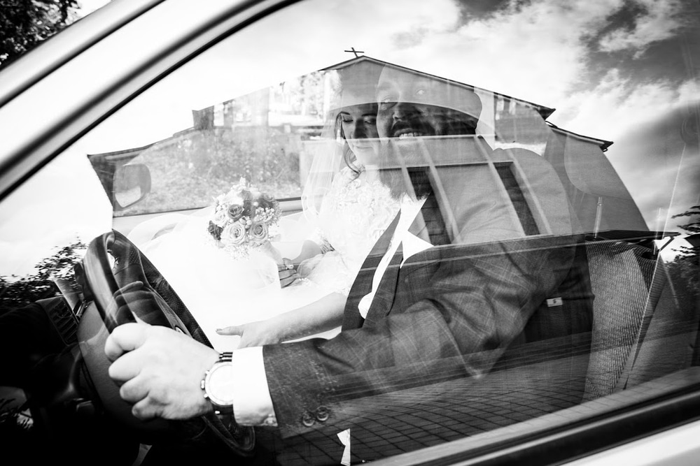 newlywed in the car in which's window reflection the church can be seen