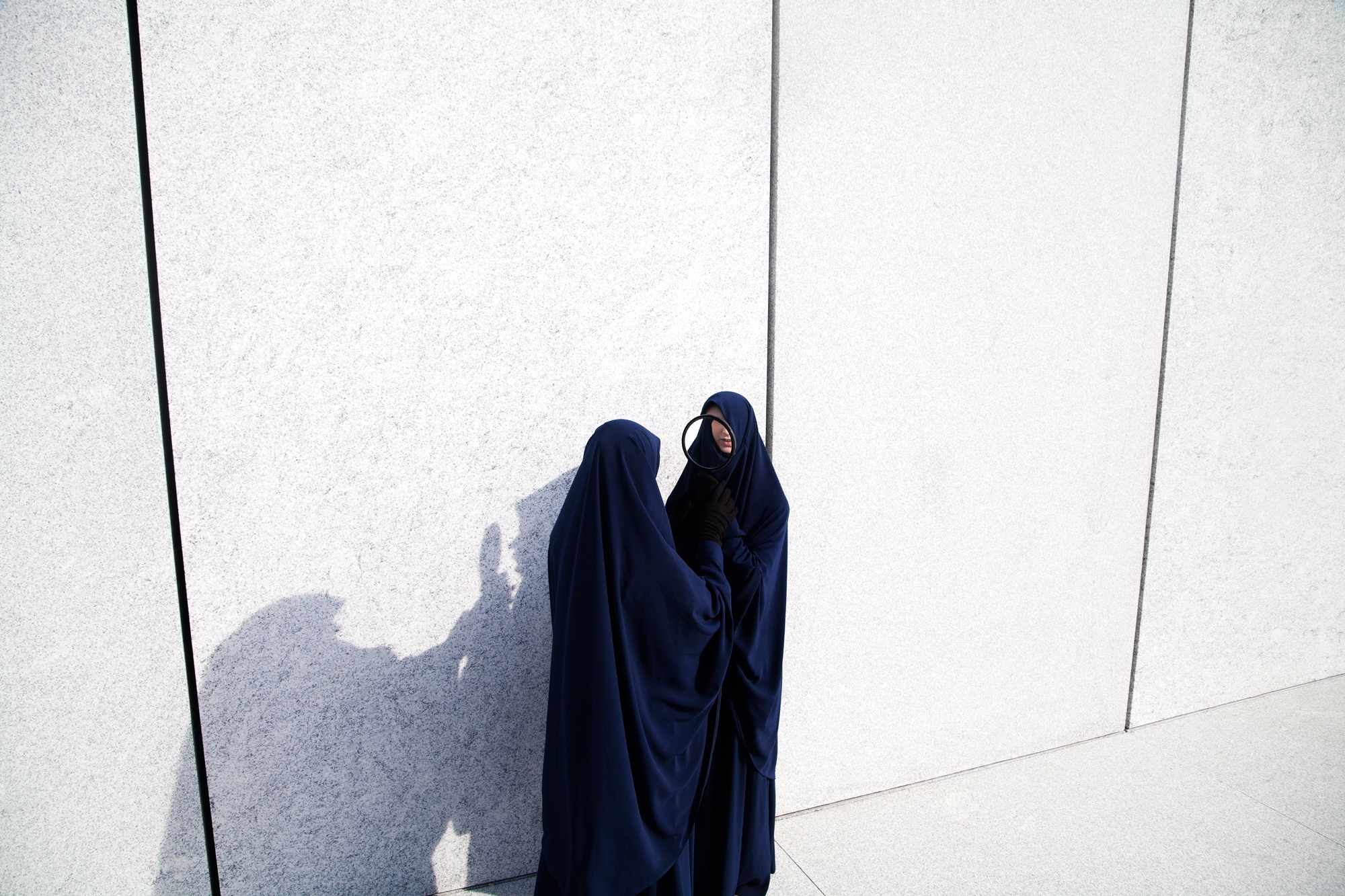 Two people in dark blue burkas stand against a giant white wall.
